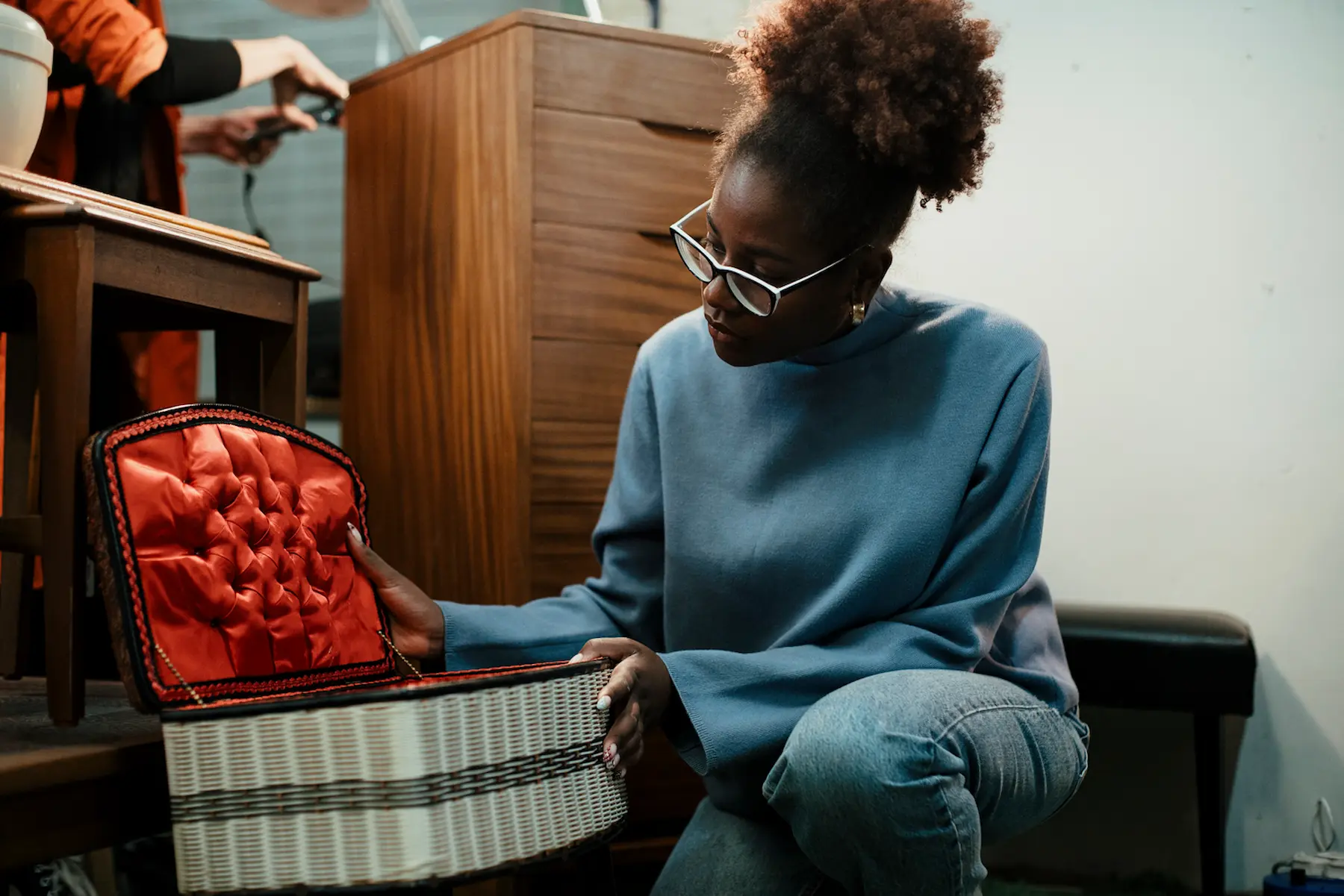 A woman in a blue sweater looks at a vintage box on the floor of a flea market