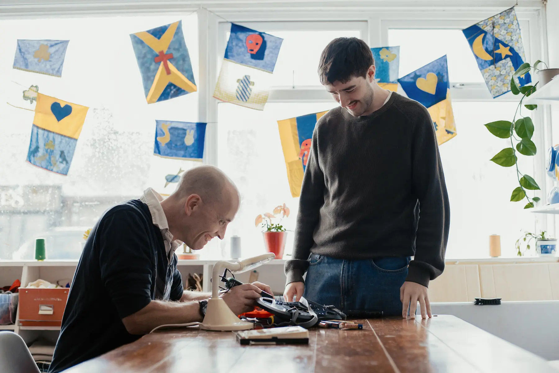 Two men repair an electronic device. The man on the left is soldering some wiring as the man on the right watches on