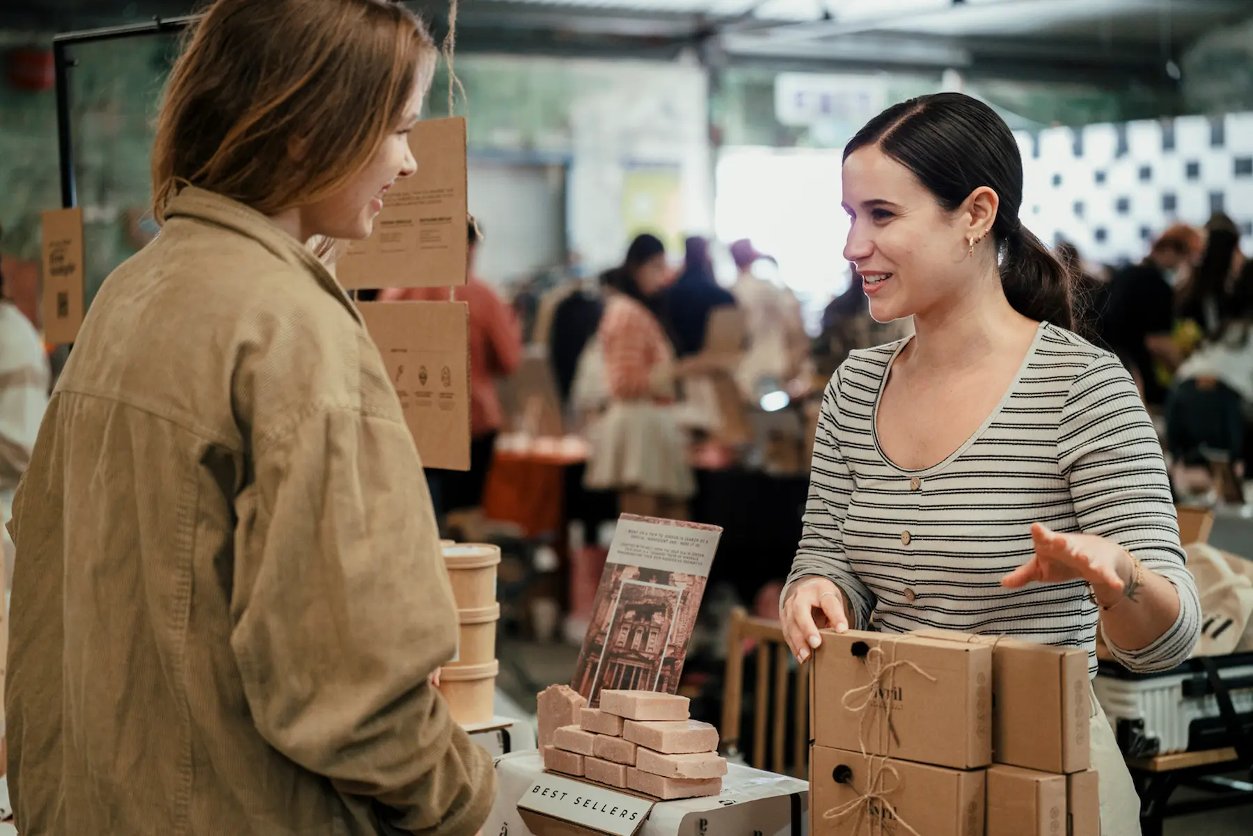 Two women chat at a flea market. A dark-haired lady on the right shows her soap products to the other woman, who is wearing a brown top
